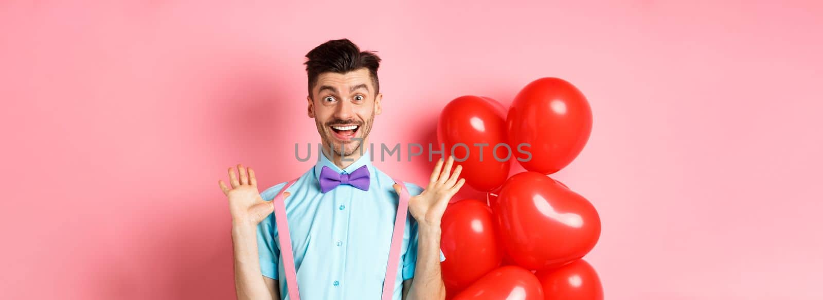 Valentines day concept. Happy young man looking surprised, raising hands up joyful, celebrating near big red hearts and pink background by Benzoix