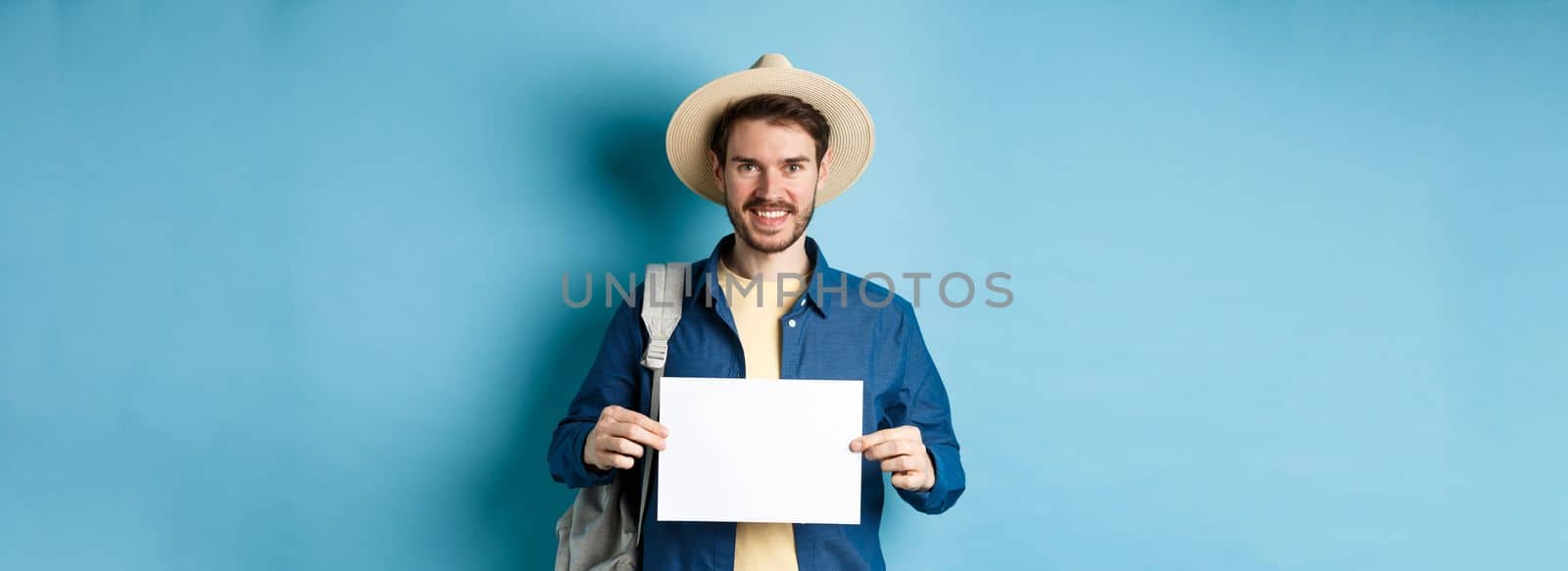 Happy tourist with backpack showing empty piece of paper, smiling at camera, standing on blue background. Concept of summer holiday and vacation.