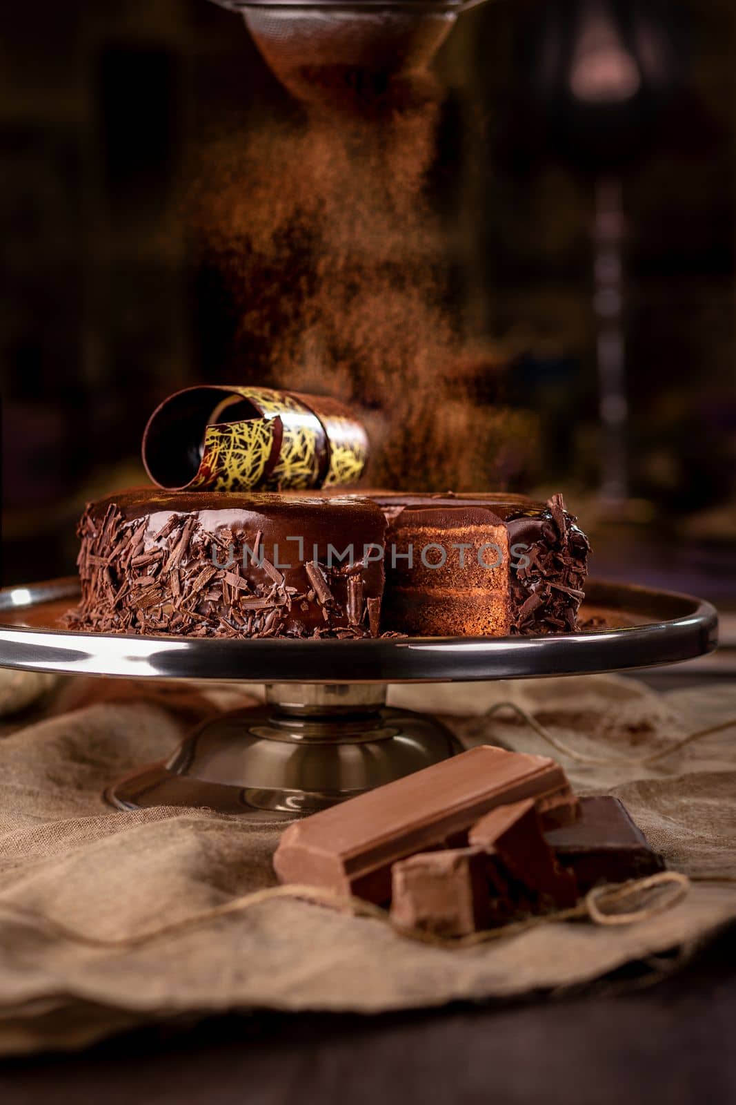 Tempting chocolate biscuit cake with chocolate buttercream layers covered with glossy ganache sprinkled with cocoa powder served on cake stand on dark background