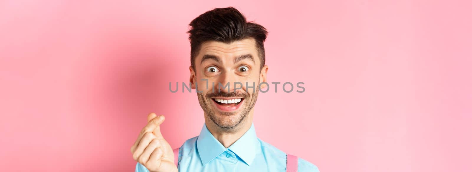 Valentines day concept. Smiling man showing finger heart and looking happy, standing over pink background.