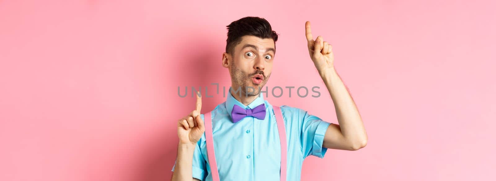 Cheerful funny man dancing in bow-tie and suspenders, pointing fingers up and looking upbeat, standing over pink background.