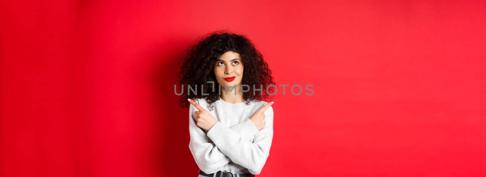 Pensive young woman making choice, pointing fingers sideways and looking at upper left corner with confident face, standing on red background.