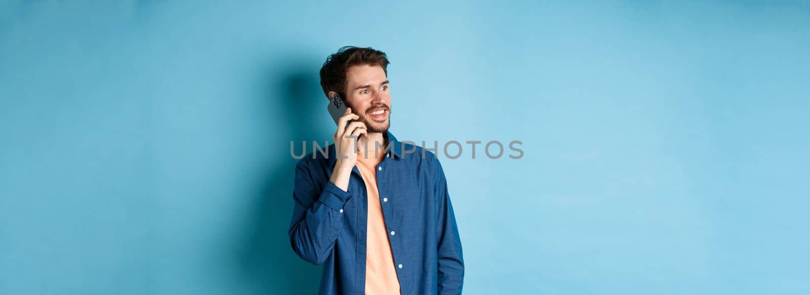 Handsome caucasian man talking on mobile phone, looking aside at empty space with carefree smile, standing on blue background by Benzoix