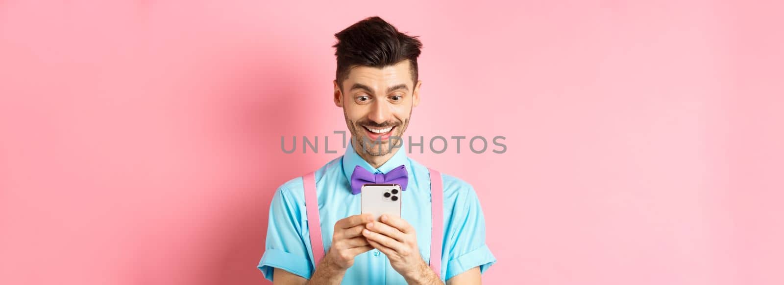 Online shopping. Happy guy looking at smartphone screen, reading message and smiling, standing on pink background.