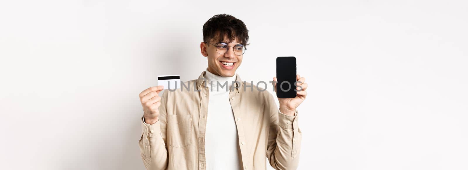 Online shopping. Handsome guy in glasses showing empty cellphone screen and credit card, smiling and looking at display pleased, standing on white background.