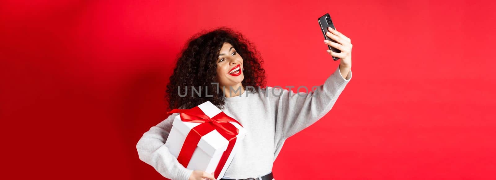 Holidays and tech concept. Happy woman taking selfie with her gift, holding present and smartphone, standing on red background.
