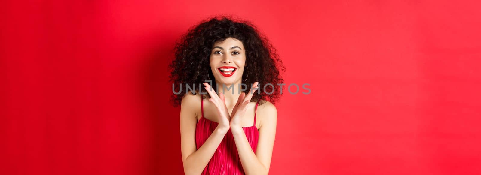 Cheerful young woman with curly hair, wearing evening dress, clap hands and smiling, praise good performance with applause, standing against red background.