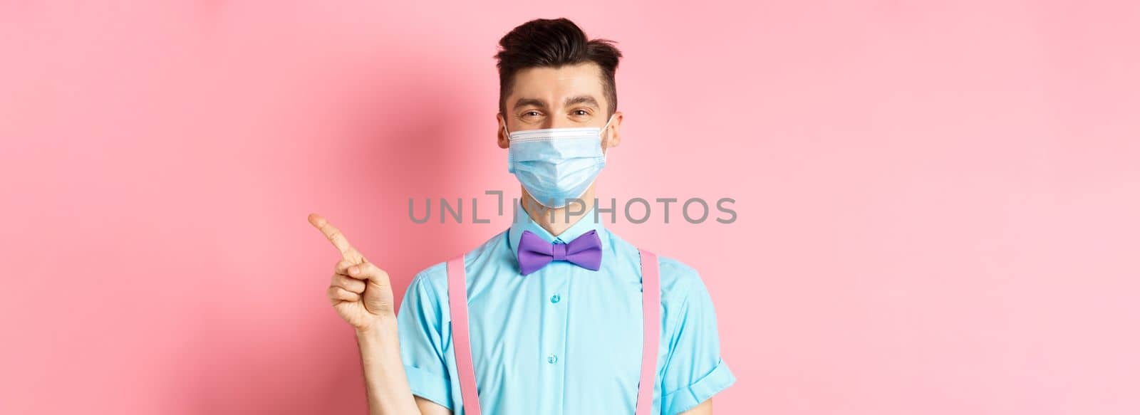 Covid-19, pandemic and health concept. Smiling caucasian man in medical mask pointing finger right, showing advertisement, standing on pink background.