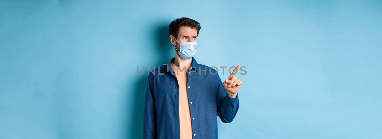 Covid-19 and healthcare concept. Confused and doubtful man in face mask frowning, pointing and looking at empty space, standing on blue background by Benzoix