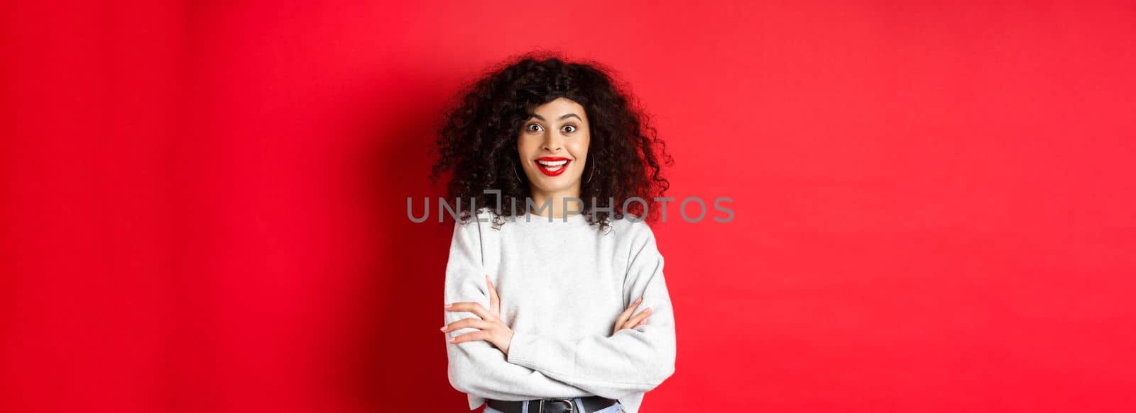 Cheerful young woman with curly hairstyle, raising eyebrows and looking surprised, hear interesting news, red background.