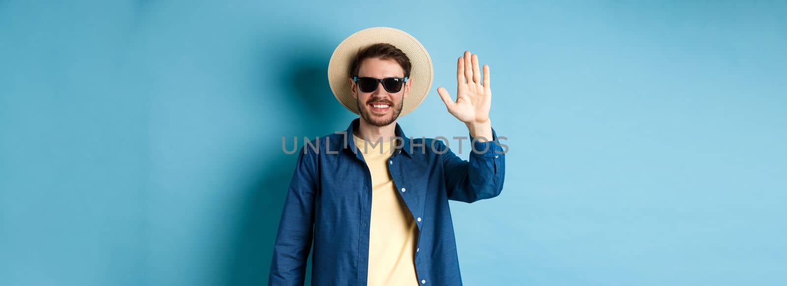 Handsome hipster on summer holiday, waiving hand and smiling, saying hello, wearing sunglasses and straw hat, blue background.