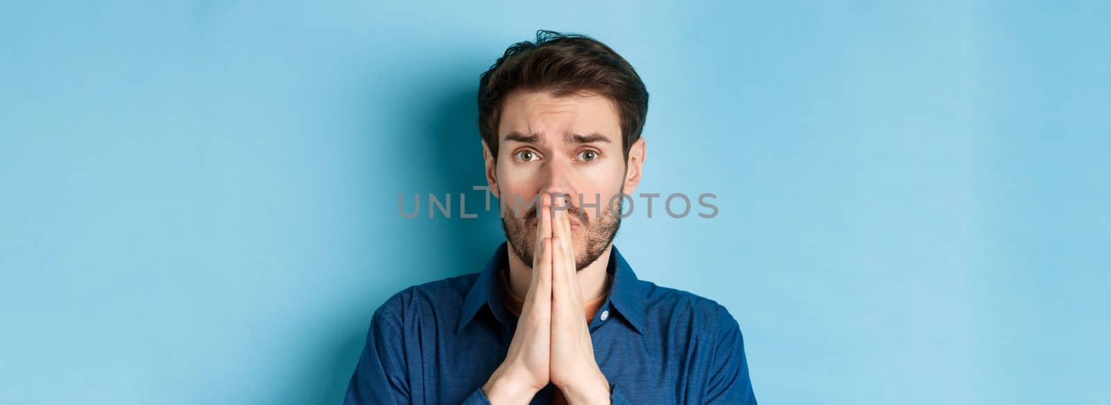 Close up of sad young man pleading, begging for help and say please, standing on blue background.