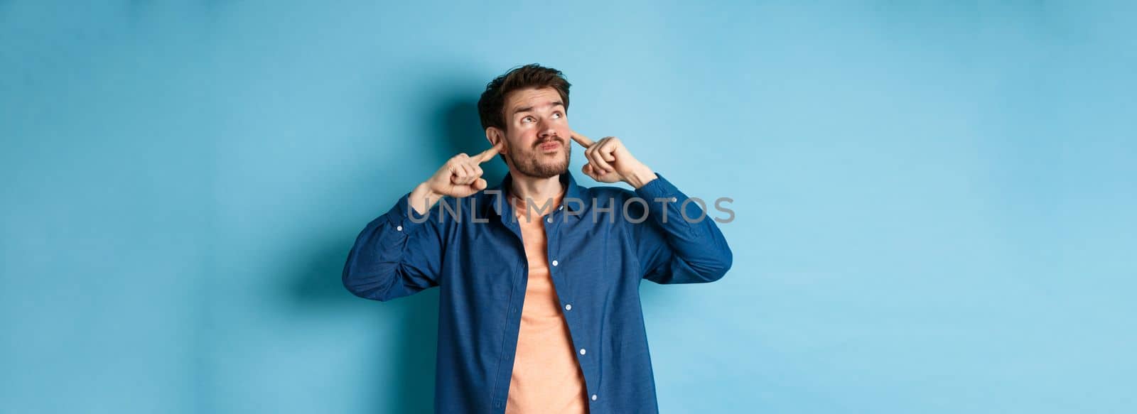 Annoyed young man block sound, shut ears with fingers and grimacing, disturbed by loud neighbours, looking up irritated, standing on blue background.