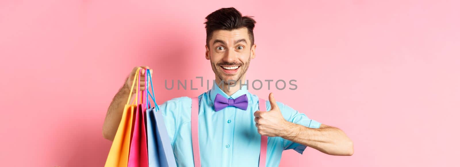 Happy male shopper showing thumbs up and shopping bags, recommending store, standing on pink background.