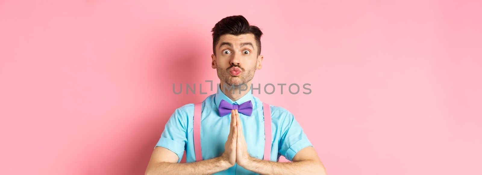 Young guy with cute face pucker lips and asking please, begging for favour, need something and lookin gpleading at camera, standing over pink background.