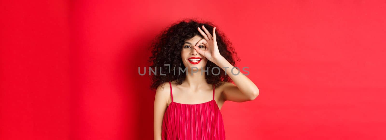 Beautiful woman look through okay sign with excitement, checking out promo offer, standing in redd against red background by Benzoix