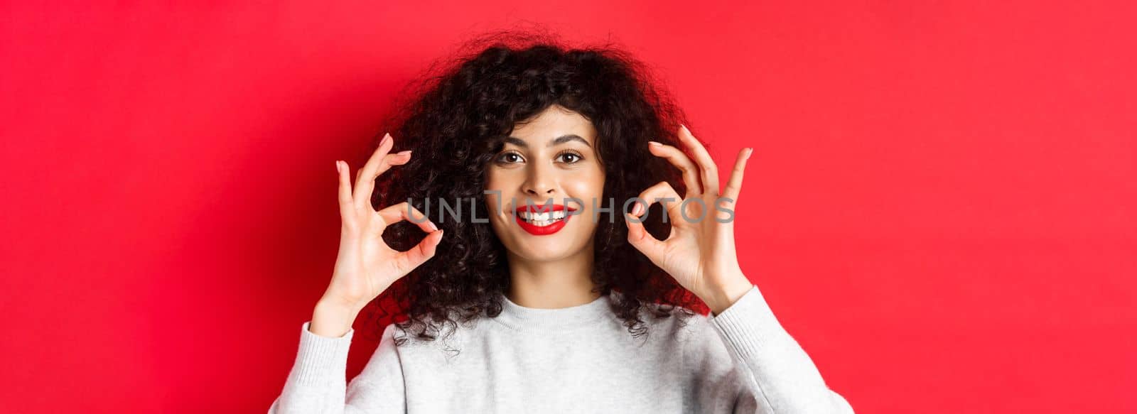 Close-up portrait of smiling woman with curly hair and red lips, showing okay gesture and looking satisfied, praise good product, standing on red background.