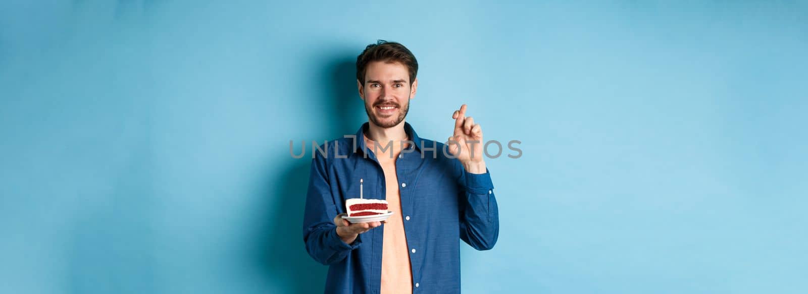 Celebration and holiday concept. Hopeful young man making wish on birthday cake, cross fingers for good luck and smiling positive, standing on blue background by Benzoix