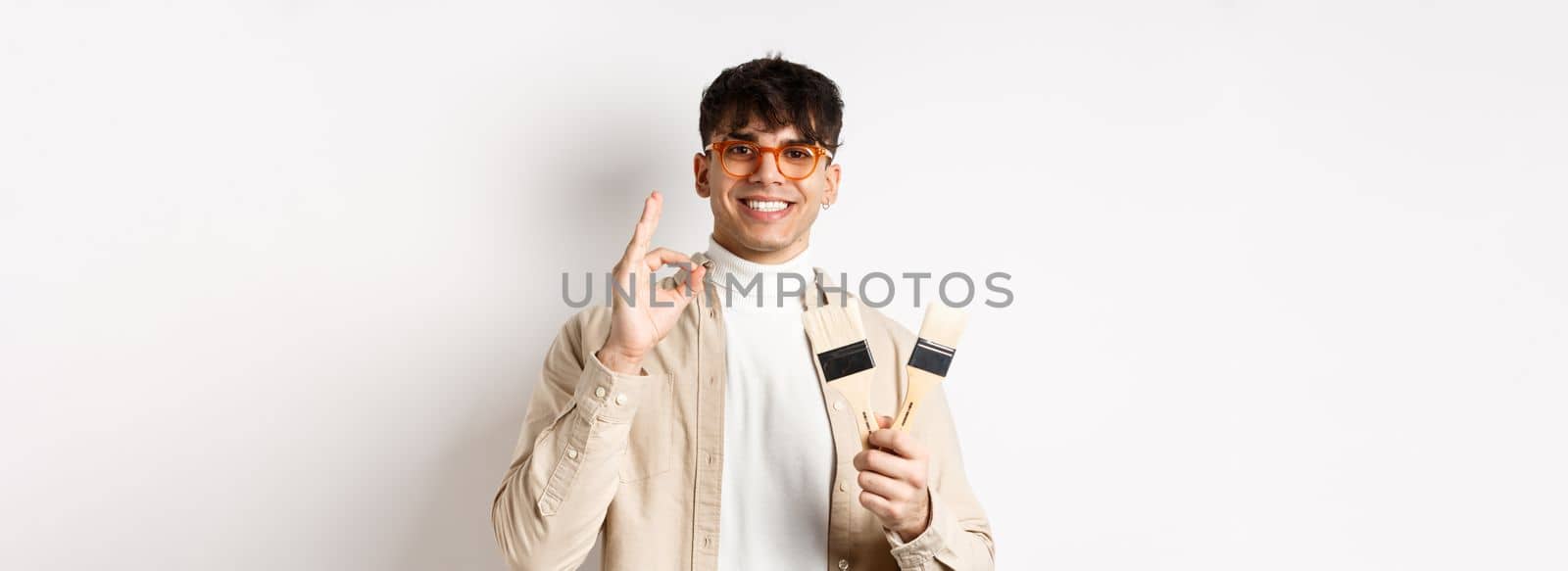 Handsome natural guy in glasses recommending store, showing painting brushes for renovation and decor, make okay sign in approval, standing on white background.