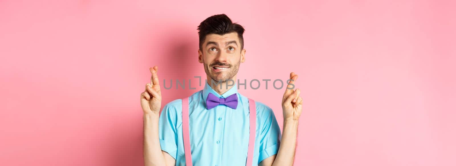 Smiling young man in bow-tie, making wish, praying and looking up, cross fingers for good luck, standing hopeful over pink background. Copy space