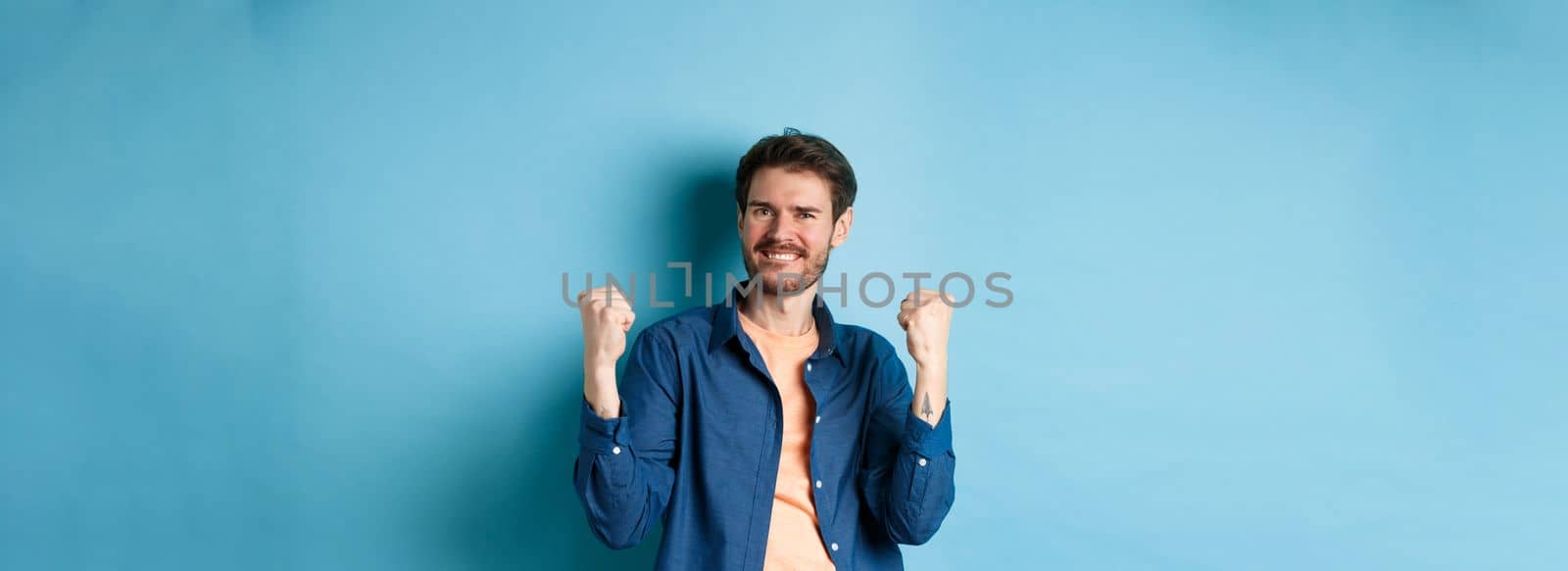 Image of happy man celebrating success, shouting yes and shaking clenched fists, achieve goal and triumphing, winning and dancing, standing on blue background.