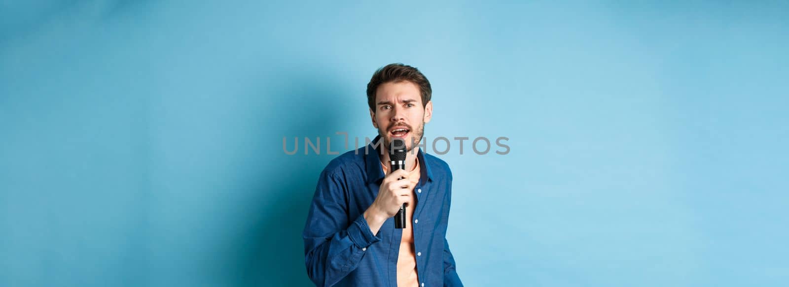 Passionate singer looking at camera, singing in microphone, playing karaoke on blue background. Copy space