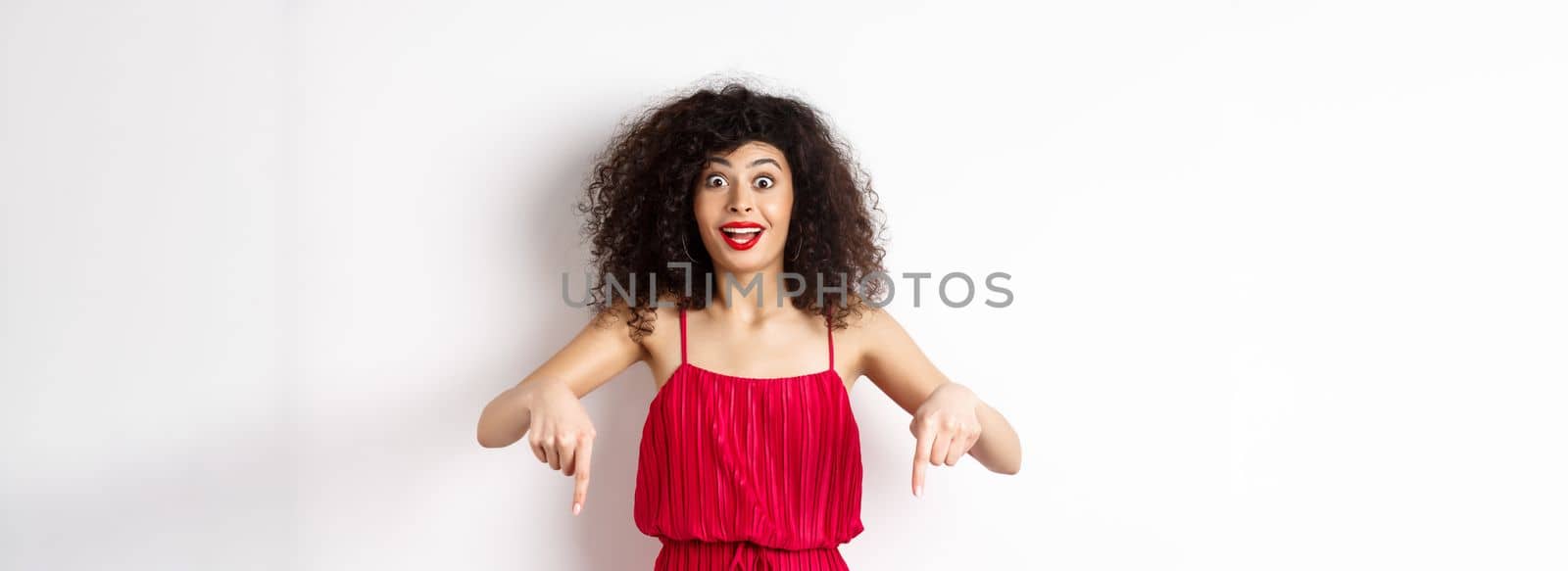 Excited woman with curly hair and red lips, wearing evening dress, gasping and pointing fingers down at super cool promo, standing on white background by Benzoix