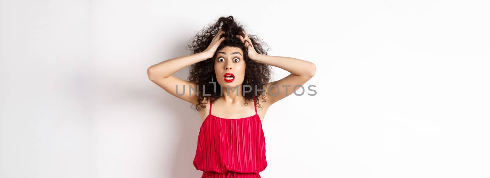 Shocked young woman holding hands on head and panicking, staring frustrated at camera, wearing red dress and makeup, white background by Benzoix