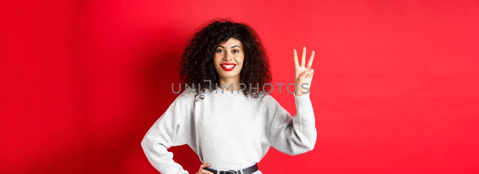 Cheerful female model showing number three and smiling, making an order, standing in sweatshirt on red background by Benzoix