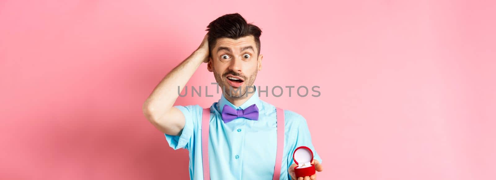 Valentines day. Confused funny guy standing with engagement ring, scratching head with unsure face, dont know what say, making proposal, standing over pink background.