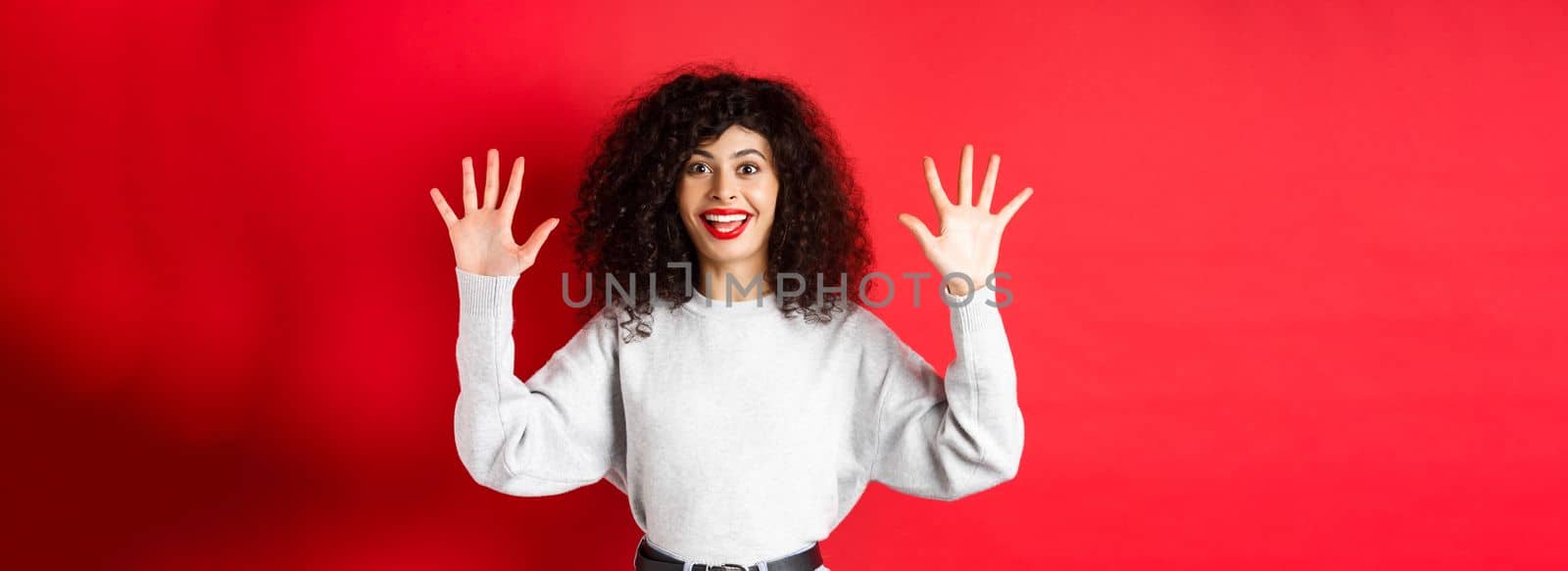 Happy beautiful woman smiling and raising hands up, showing number ten, ordern dozen of something, standing in sweatshirt on red background.