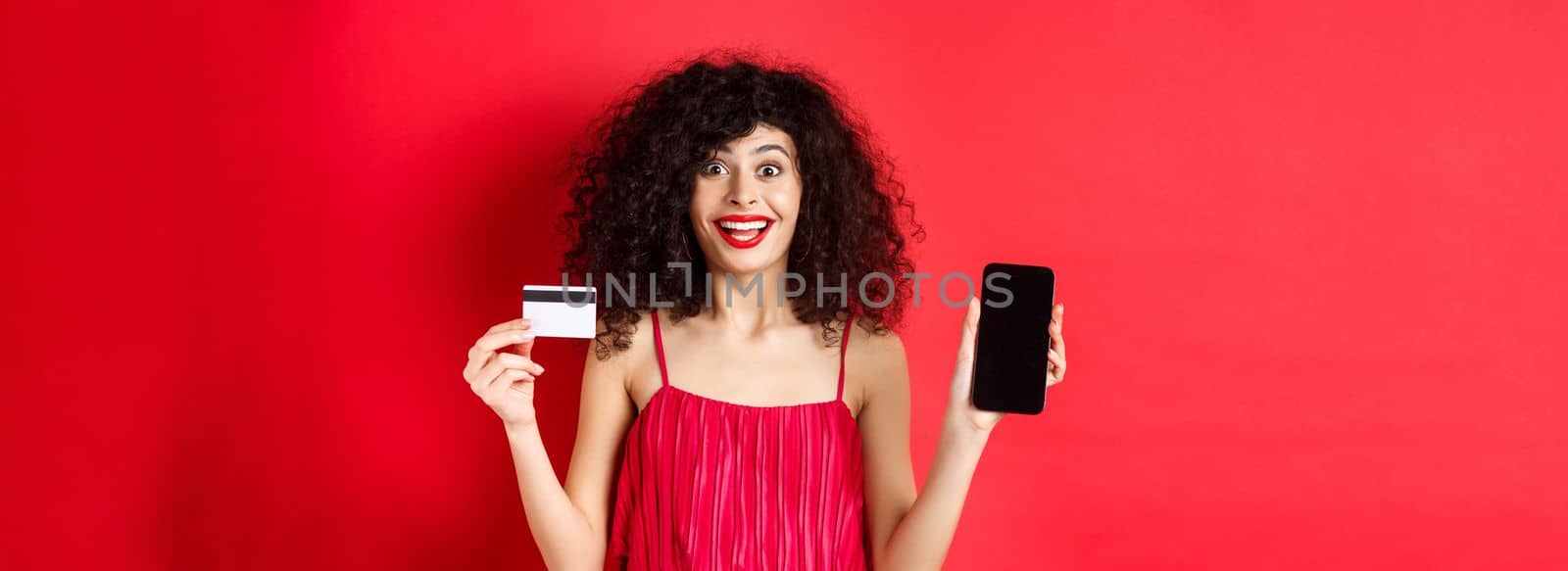 Excited lady in red dress showing plastic credit card and smartphone screen, smiling amazed, standing on studio background by Benzoix