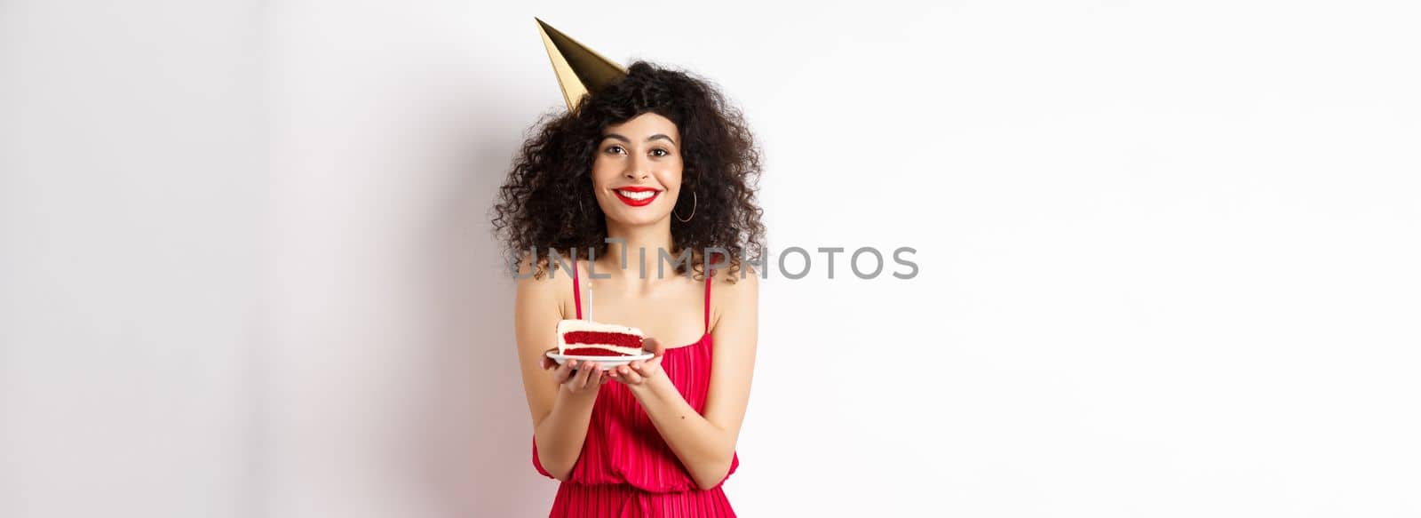 Beautiful woman in red dress, wearing party hat and celebrating birthday, holding b-day cake and making wish, smiling at camera, standing on white background.