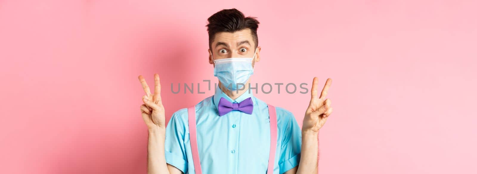 Coronavirus, healthcare and quarantine concept. Funny guy in bow-tie and face mask showing peace, victory signs and looking happy at camera, standing on pink background by Benzoix