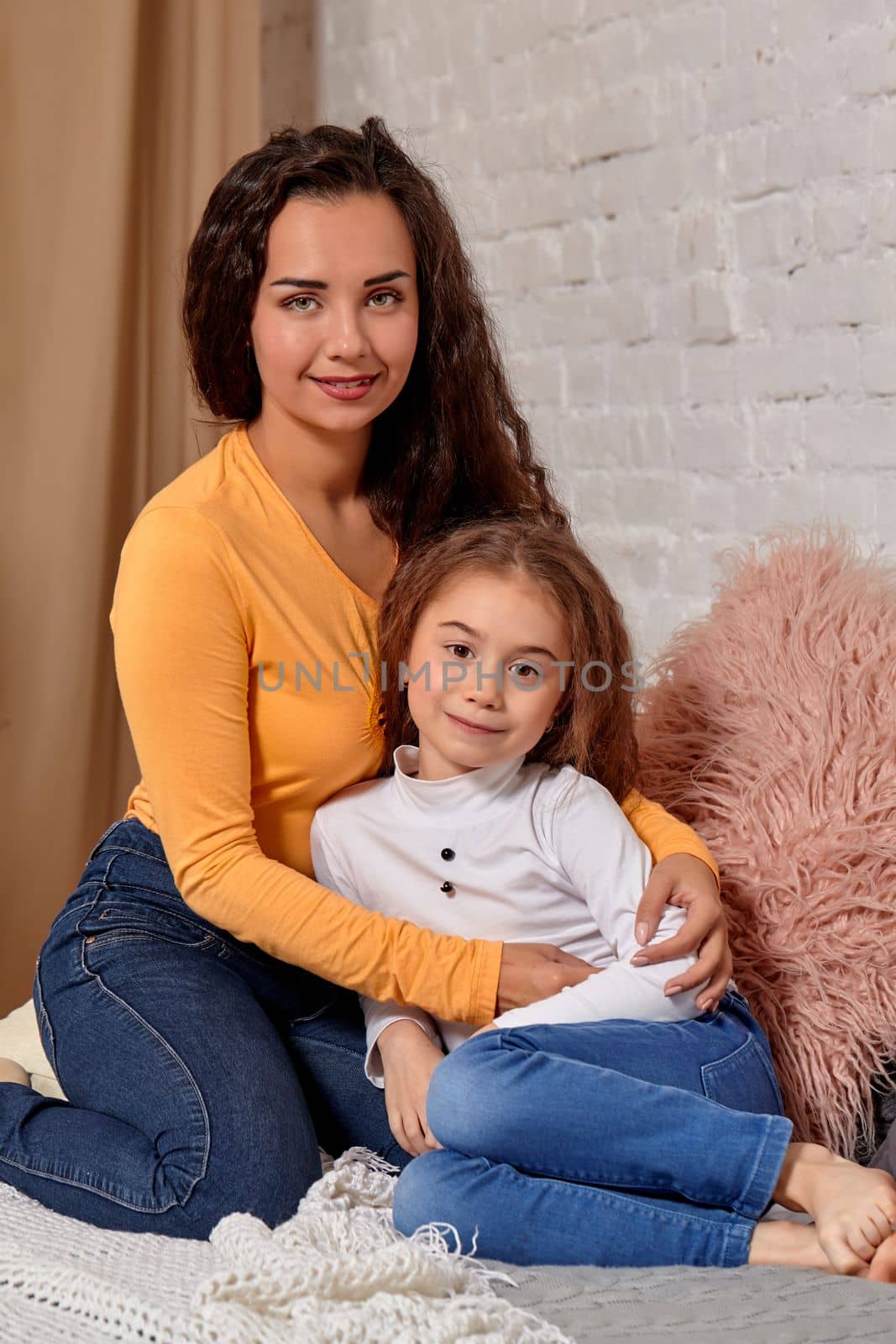 Love of young mother and daughter. They sit on the bed at home and have fun, mother hugs daughter