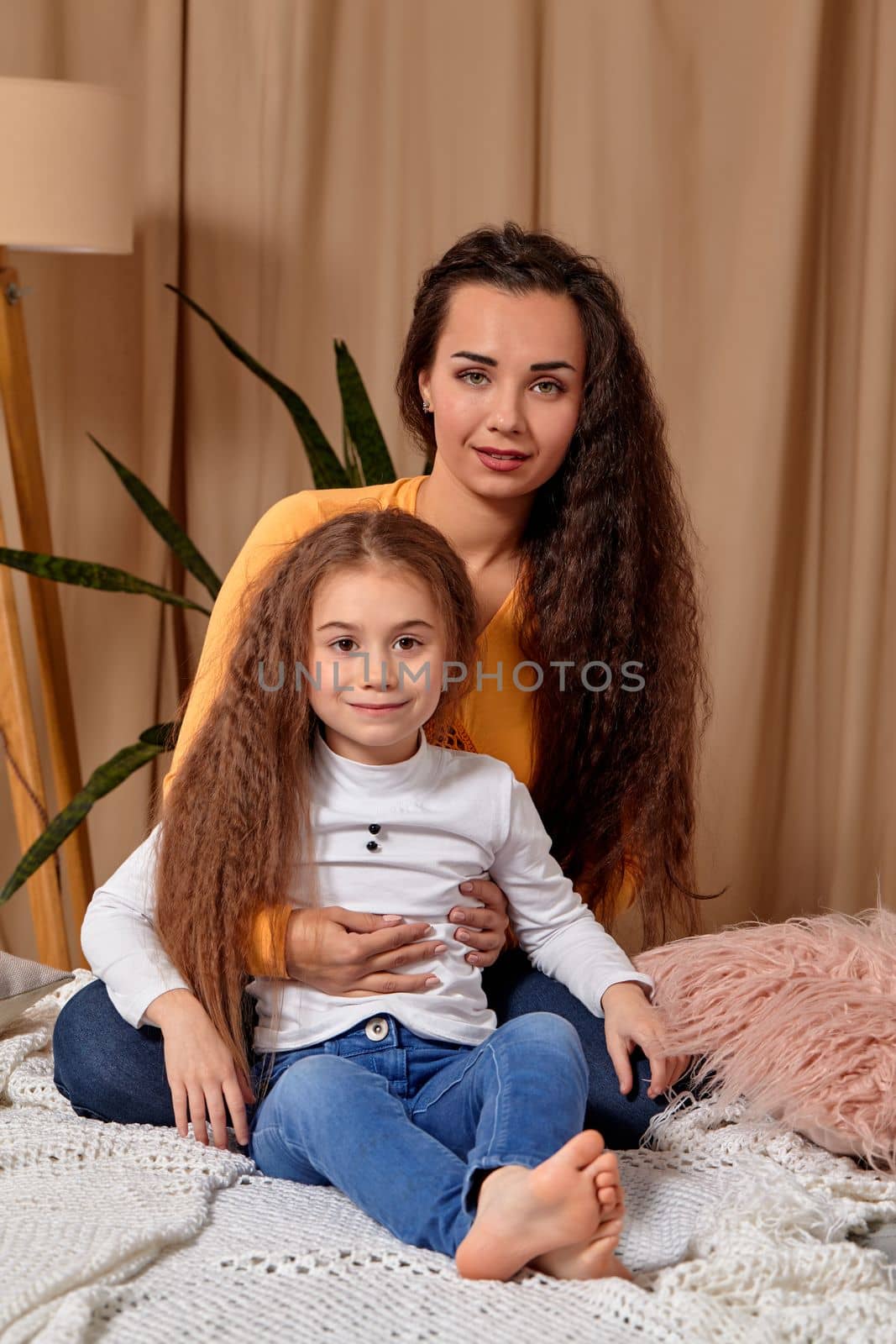 Love of young mother and daughter. They sit on the bed at home and have fun, mother hugs daughter