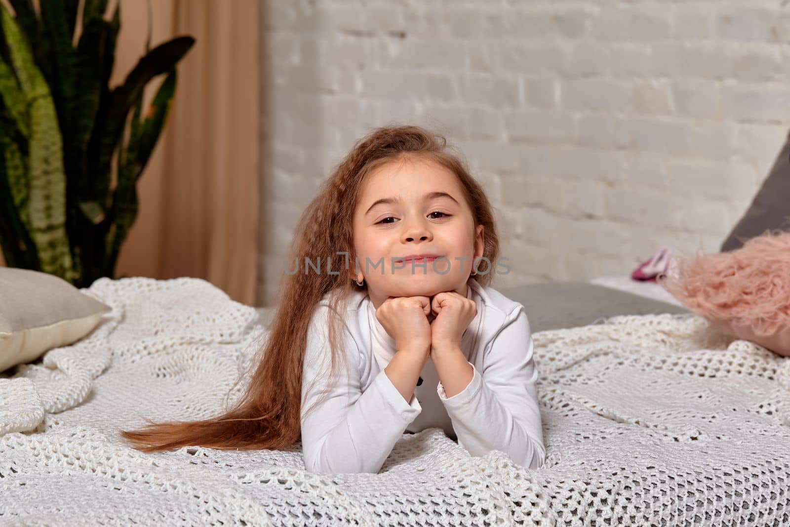 A little girl lies on the bed in the room. She posing before camera and looks happy
