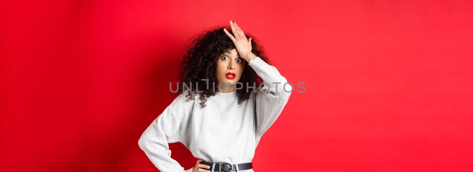 Annoyed and tired young woman slap forehead, making facepalm and stare at camera shocked, standing bothered against red background.