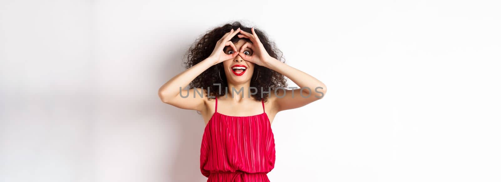 Impressed young woman seeing cool thing, look through hand binoculars and say wow, standing amazed at white background by Benzoix