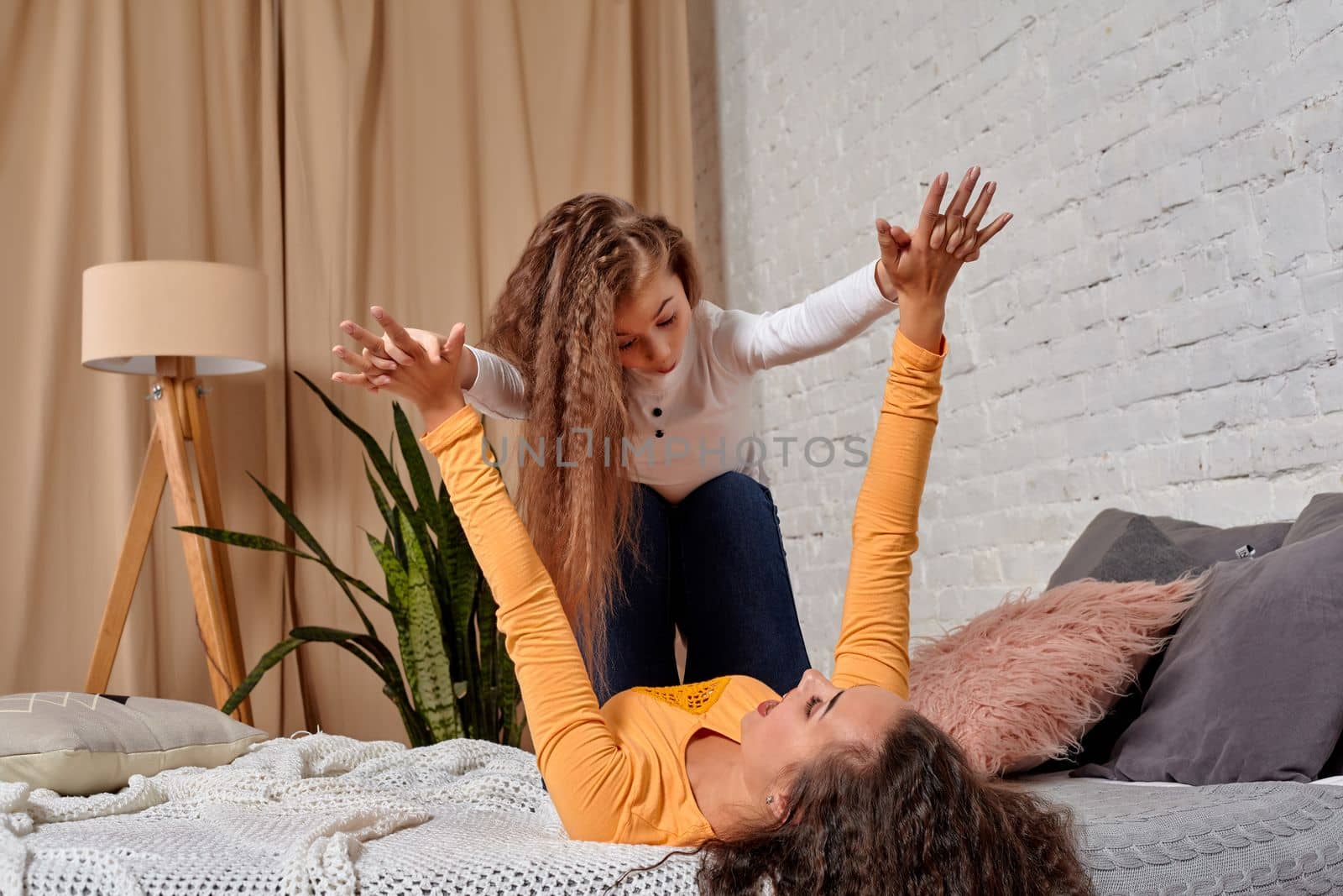 Love of young mother and daughter. They lie on the bed at home and have fun, they are fooling around, mother is holding her daughter on top of her legs