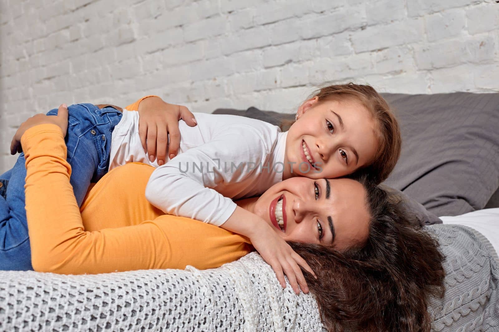 Love of young mother and daughter. They lie on the bed at home and have fun, daughter lies on top of mom, they hug each other