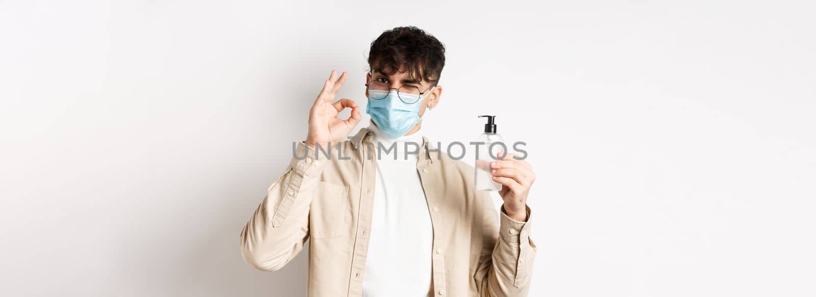 Health, covid and quarantine concept. Portrait of natural guy in glasses and face mask showing bottle of good hand sanitizer, make okay sign, recommending product, white background.