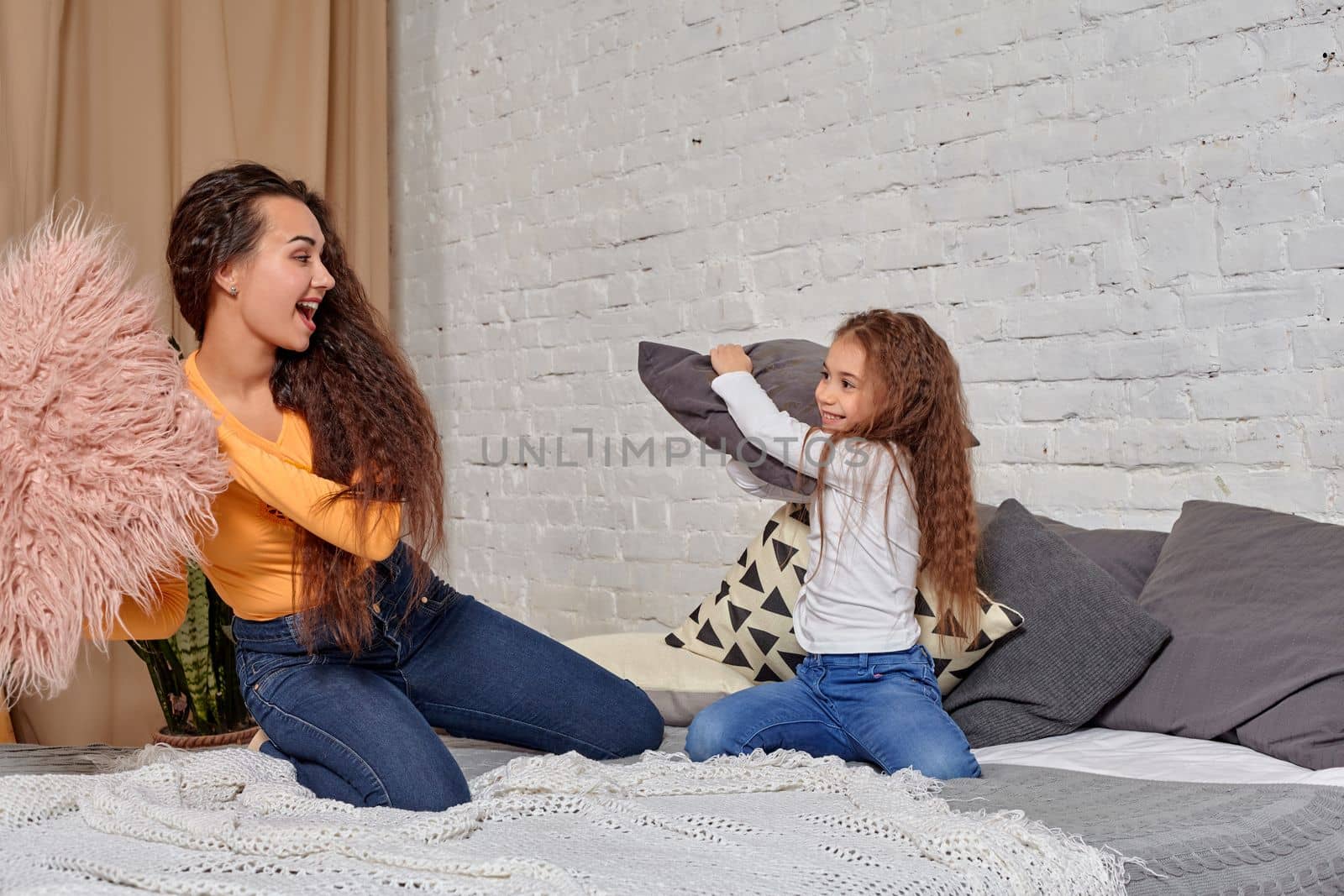 mom and daughter sit on bed, fooling around with pillow fights, they have fun, they looks happy