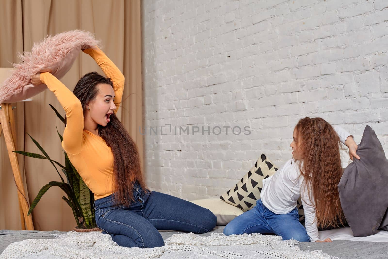 mom and daughter sit on bed, fooling around with pillow fights, they have fun, they looks happy