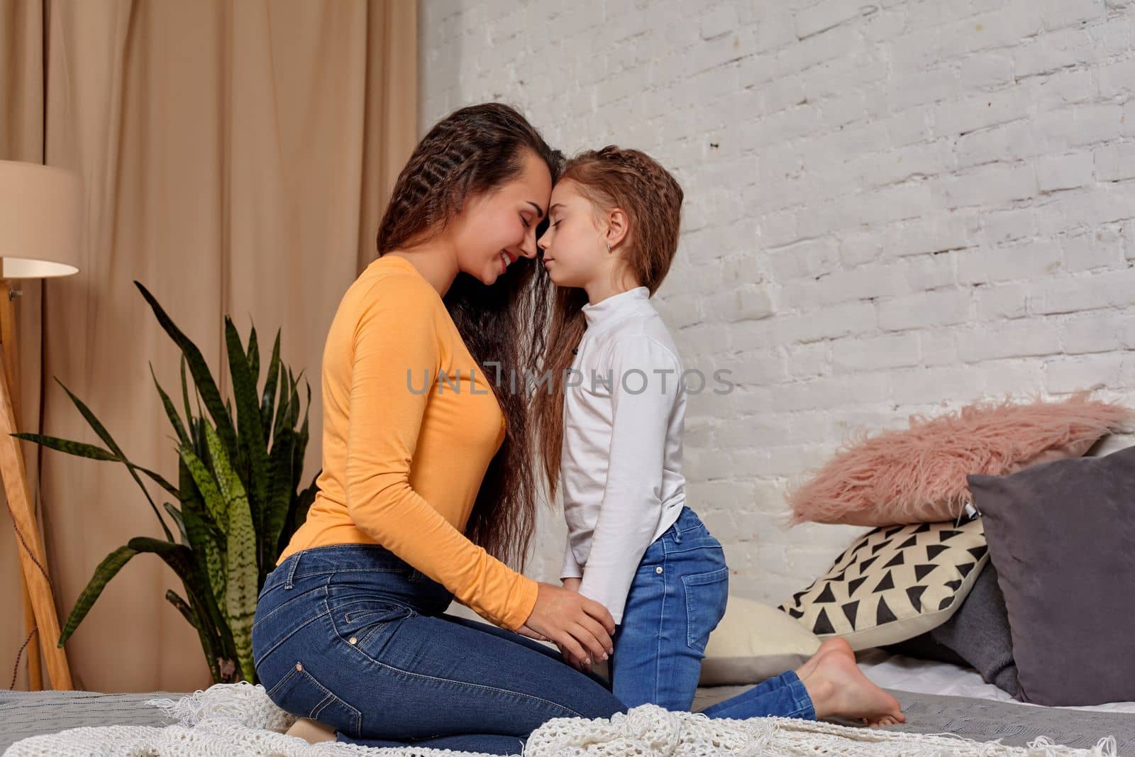 Love of young mother and daughter. They sit on the bed opposite each other and show emotions, touching their foreheads, and holding hands