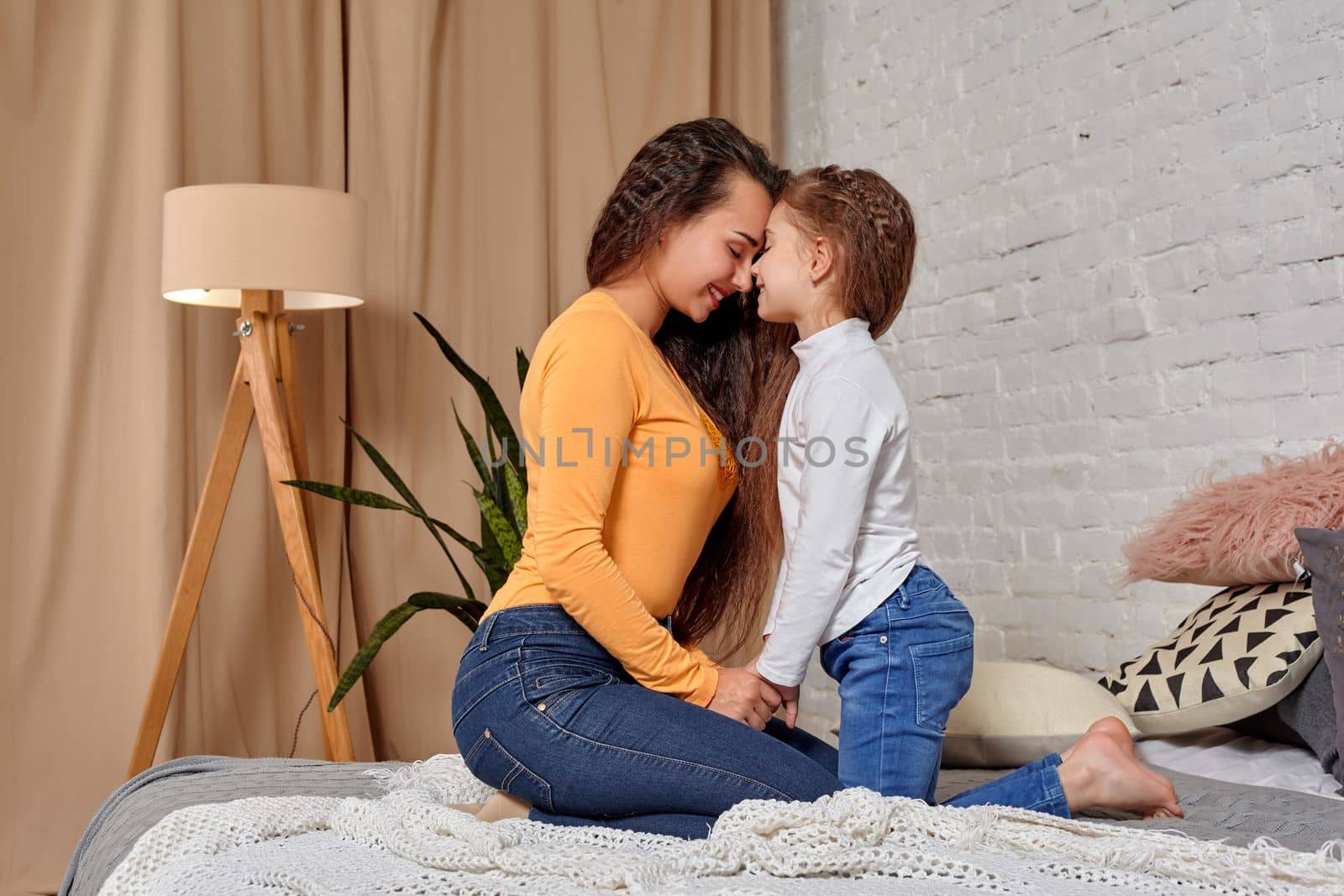 Love of young mother and daughter. They sit on the bed opposite each other and show emotions, touching their foreheads, and holding hands