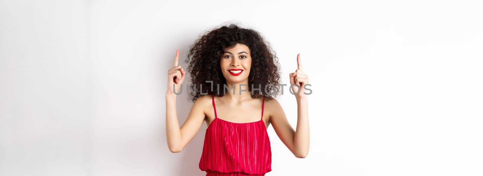 Smiling woman in elegant red dress and makeup, pointing fingers up and showing promo offer on valentines day, standing over white background by Benzoix