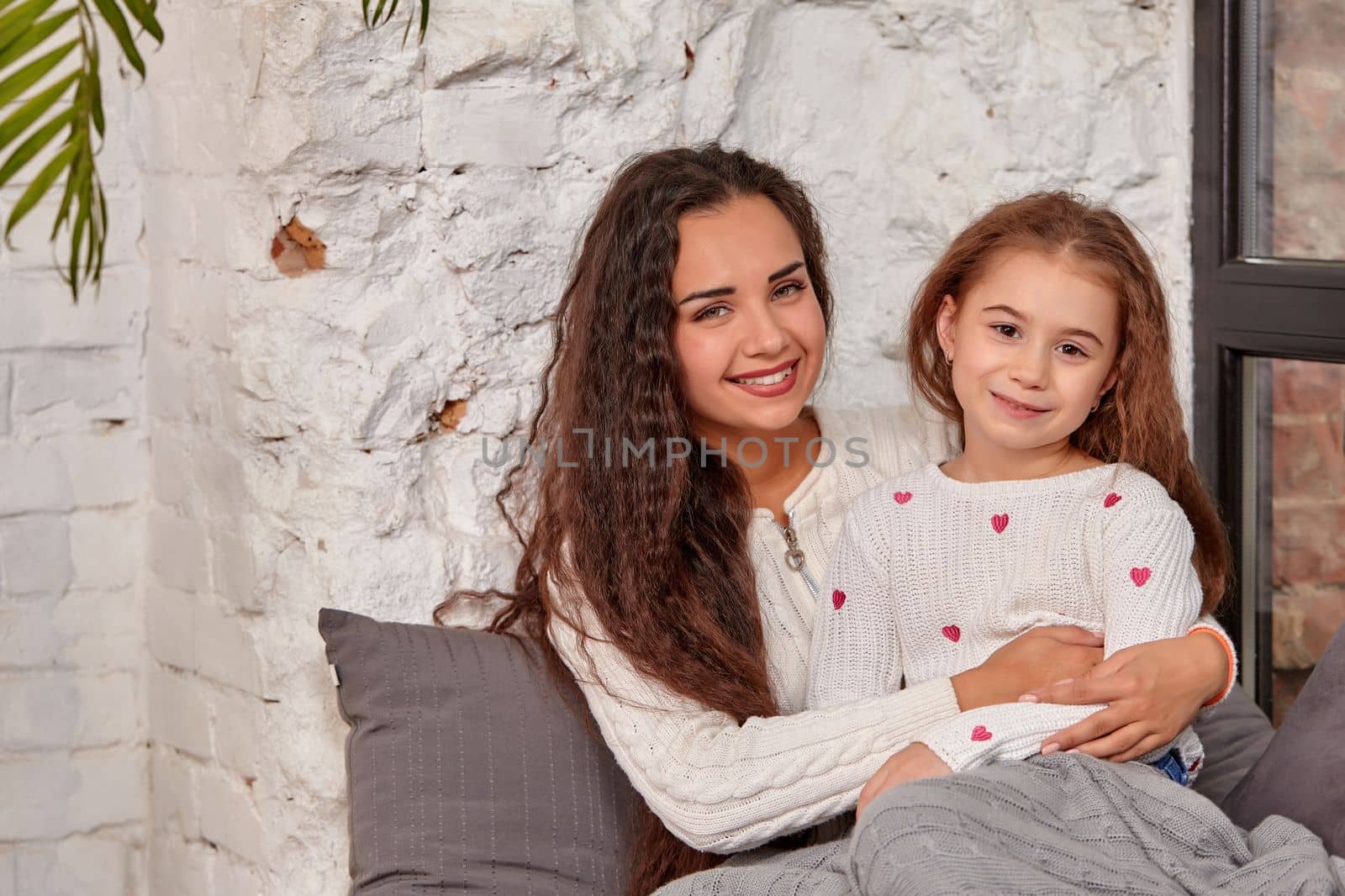 Mother and daughter sitting on sill near window in room. They show emotions and have fun