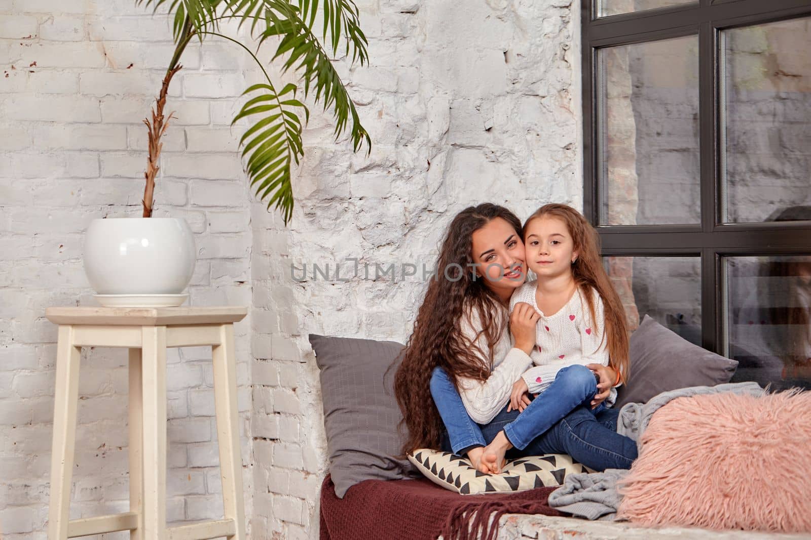 Mother and daughter sitting on sill near window in room. They show emotions and have fun