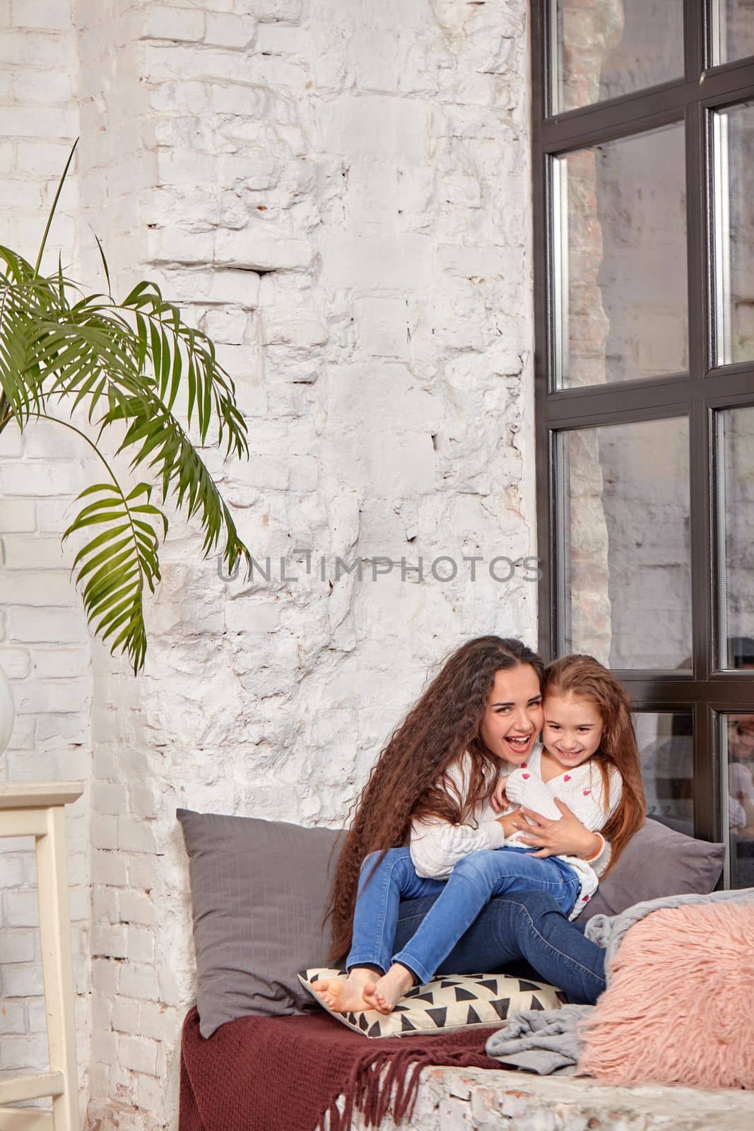 Mother and daughter sitting on sill near window in room. They show emotions and have fun